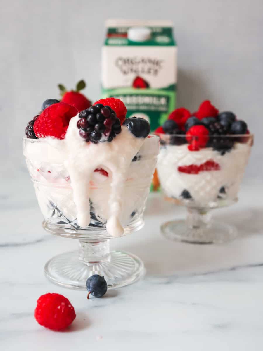 The image depicts a transparent vintage glass ice cream dish filled with layers of colorful ingredients, showcasing a Homemade Greek Yogurt parfait. The base layer consists of creamy Greek yogurt, thick and rich in texture, filling the bottom of the glass. On top of the yogurt are alternating layers of vibrant fruits, including ripe raspberries and juicy blueberries, adding bursts of color and sweetness to the parfait. The parfait is topped with more fruit. The arrangement is visually appealing, with the various layers creating a beautiful and delicious display. A half gallon of Organic Valley milk is displayed in the background.