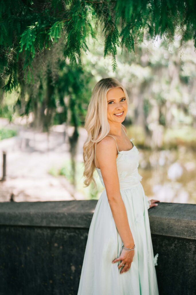 A woman with long, wavy blonde hair smiles warmly at the camera while wearing a light green dress. She's positioned in front of a lush, green outdoor background with trees and foliage creating a natural, softly blurred backdrop. The lighting appears soft and natural, giving the portrait a bright, cheerful atmosphere.