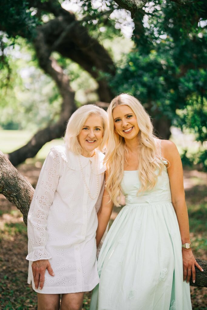 Two women with blonde hair pose together outdoors under large trees with dappled sunlight filtering through the foliage. The woman on the left wears a white eyelet lace dress with long sleeves, while the woman on the right wears a a light green dress and a gold watch. Both are smiling warmly at the camera in City Park in New Orleans.