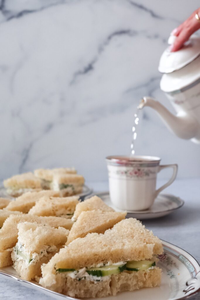 Hot tea pours from a white teapot into a delicate floral china teacup, with triangular cucumber tea sandwiches arranged on a matching plate in the foreground, set against a marble surface for an elegant afternoon tea service.