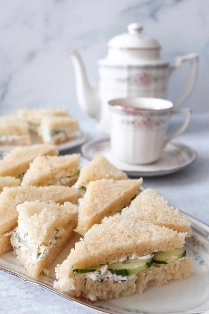 Delicate cucumber tea sandwiches with cream cheese and herbs on white bread, cut into triangles and arranged on a floral china plate, served alongside a matching teapot and teacup for an elegant afternoon tea setting.