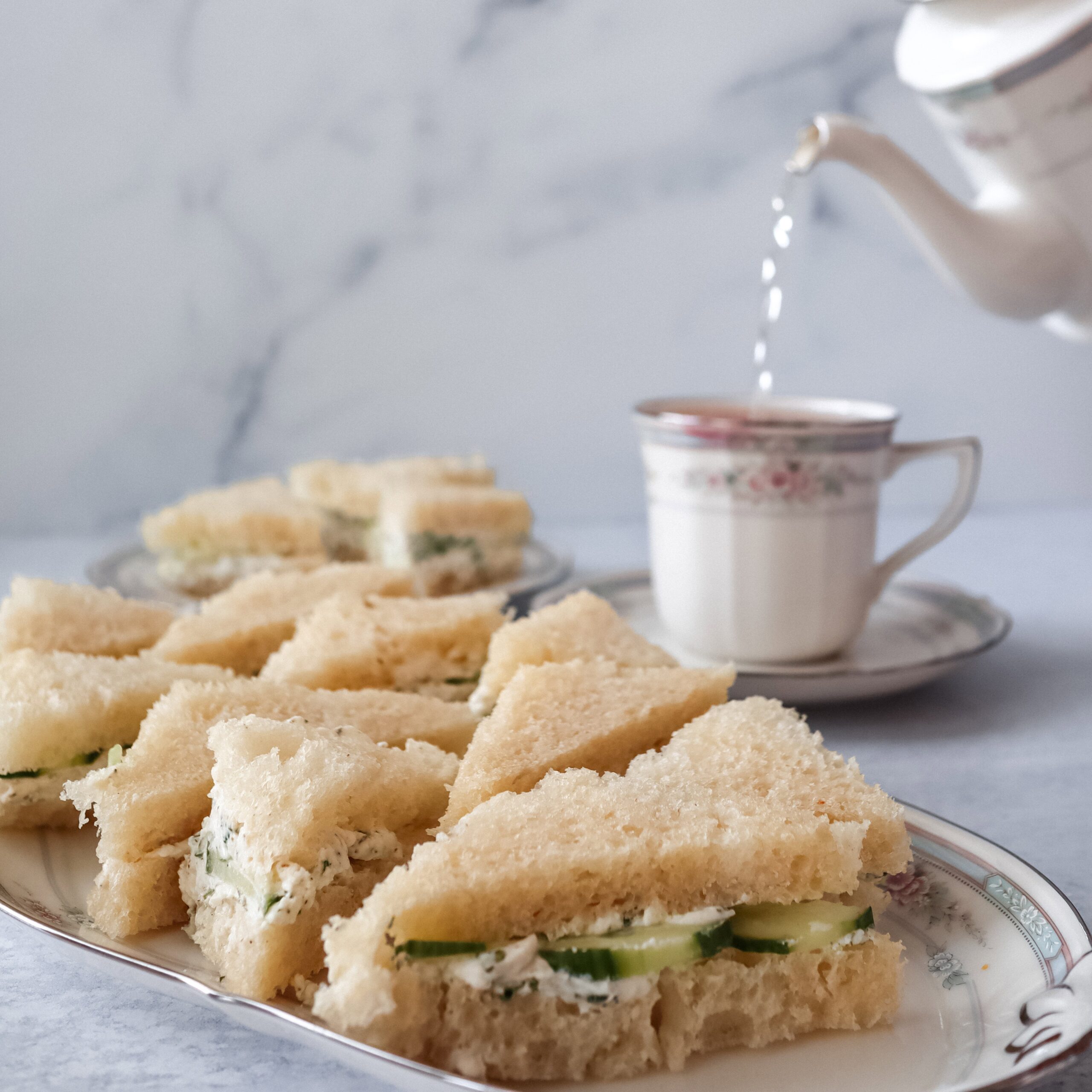 Delicate cucumber tea sandwiches with cream cheese and herbs on white bread, cut into triangles and arranged on a floral china plate, served alongside a matching teapot and teacup for an elegant afternoon tea setting.