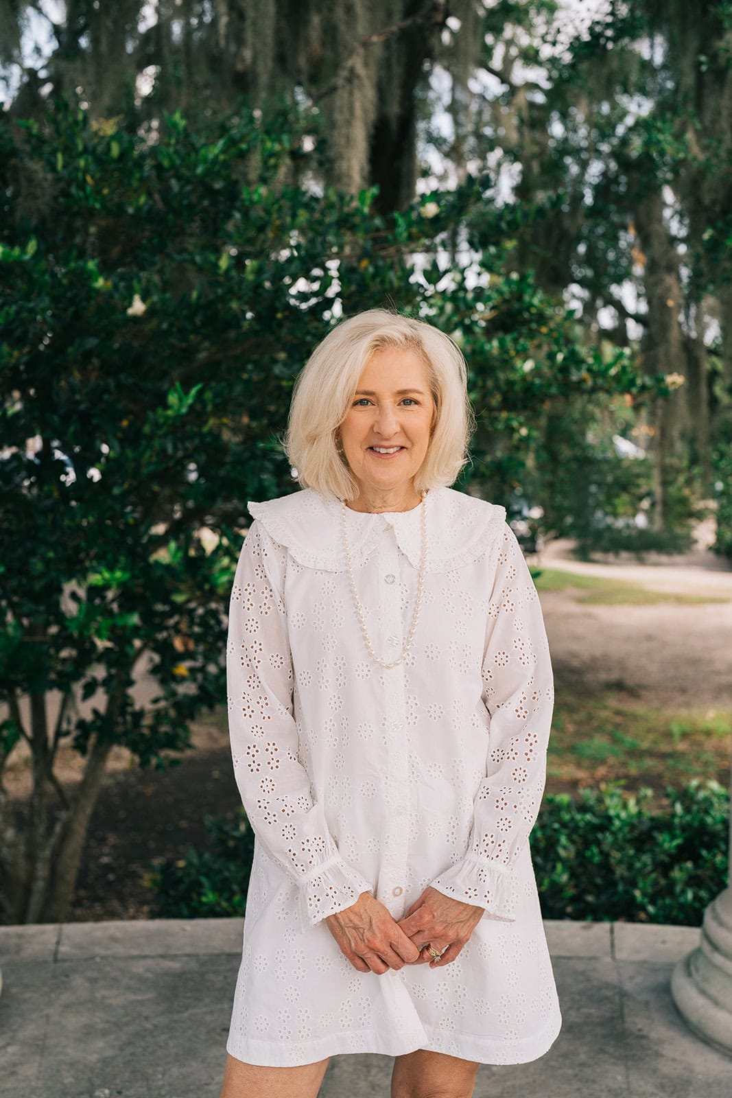 A woman with short blonde hair wearing a white eyelet lace dress with long sleeves and a collar, standing outdoors in front of lush green foliage and trees. She is smiling at the camera with her hands clasped in front of her at park.