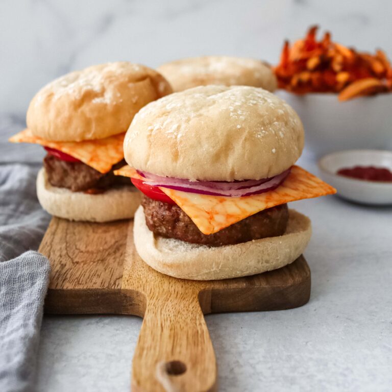 Three Turkey Burgers on soft white buns sit on a wooden cutting board. Each burger features a turkey patty topped with melted orange cheese, red onion slices, and tomato. Sweet potato fries are visible in the background along with a small bowl of ketchup.