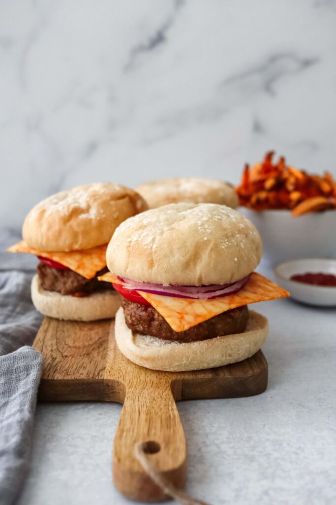Three Turkey Burgers on soft white buns sit on a wooden cutting board. Each burger features a turkey patty topped with melted orange cheese, red onion slices, and tomato. Sweet potato fries are visible in the background along with a small bowl of ketchup.