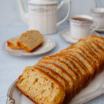Sliced lemon poppy seed bread arranged on a china platter with a pot of tea and teacups behind it