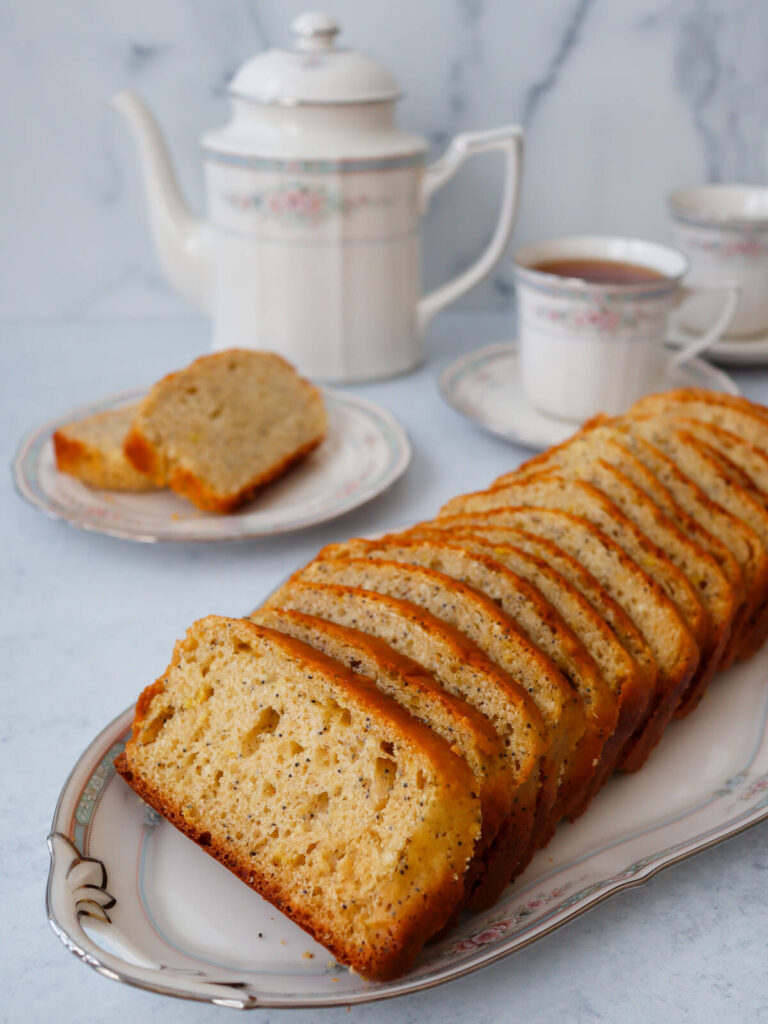 Sliced lemon poppy seed bread arranged on a china platter with a pot of tea and teacups behind it